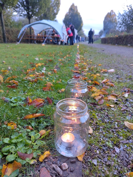Teelichter im Glas auf dem Weg im Kleingarten. Dahinter stehen Pavillons und Menschen.